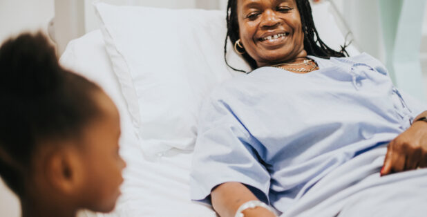 A smiling female patient lays in a hospital bed and holds the hand of her young daughter