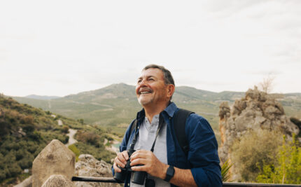 An older man looks at the view through binoculars while standing on a scenic hilltop.